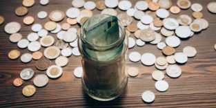 Münzen liegen auf einem Tisch, auf dem außerdem ein Glas mit Geldscheinen steht