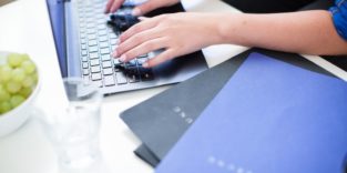 zwei Hände an einem Laptop, daneben liegen Bewerbungsmappen