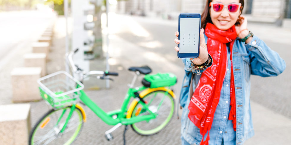 Grünes Leihrad steht hinter einer jungen Frau, die ihr Smartphone in die Kamera hält