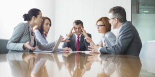 Fünf Kollegen sitzen am Besprechungstisch, vier scheinen sich zu streiten