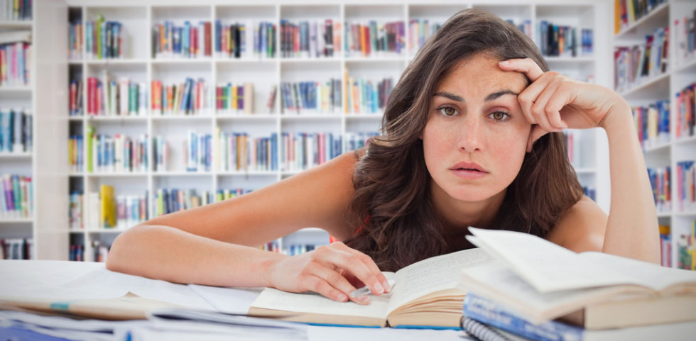 Junge Frau in einer Bibliothek hängt über einem Buch und schaut genervt in die Kamera