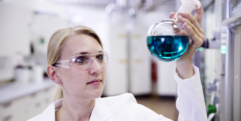 Eine Wissenschaftlerin hält einen Erlenmeyerkolben mit einer blau-durchsichtigen Flüssigkeit hoch