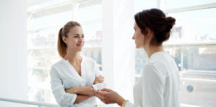 Jobsharing-Symbolbild. Zwei Geschäftsfrauen unterhalten sich vor Glasfassade