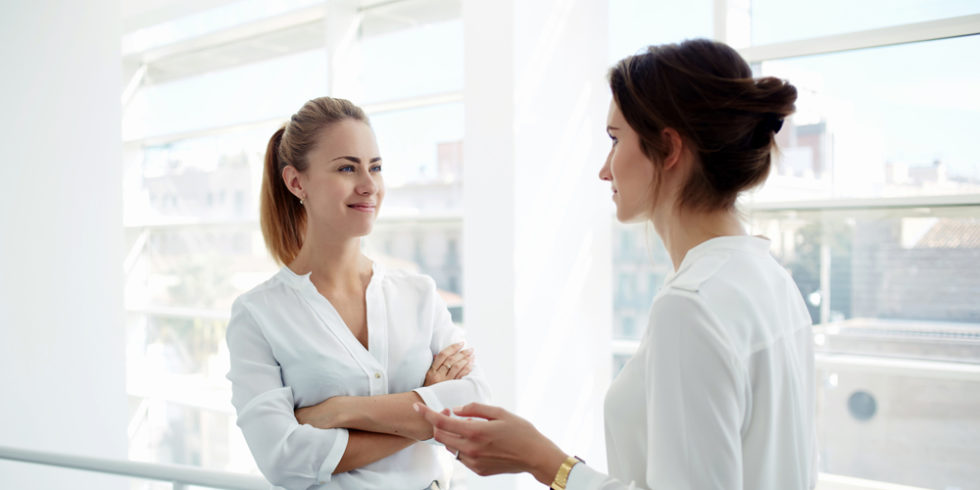 Jobsharing-Symbolbild. Zwei Geschäftsfrauen unterhalten sich vor Glasfassade