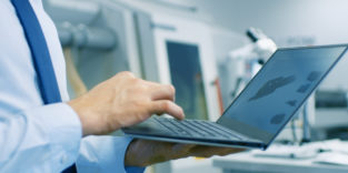 Digitales Arbeiten. Ein Mann steht mit Laptop in der Hand im Raum, dahinter eine industrieroboter