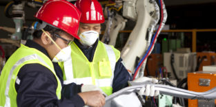 zwei Männer mit Warnwesten, Helmen und Mundschutz in der Fabrik. Im Hintergrund eine Maschine