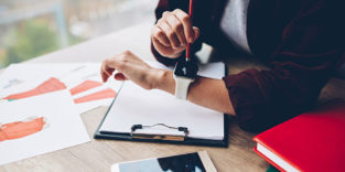 Zeiterfassung: Blick auf Frauenhände mit Smartwatch, daneben liegen auf dem Tisch Tablet und Notizpapier