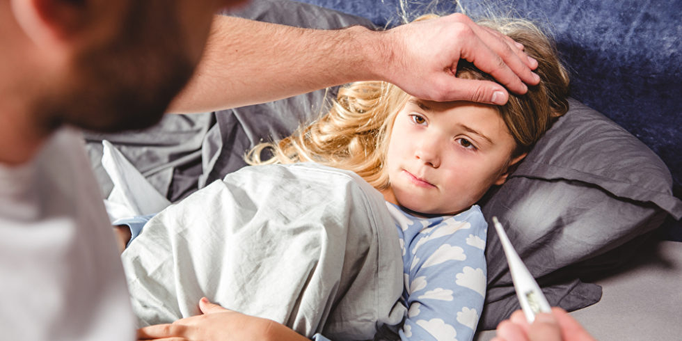 Mädchen liegt im Bett, Vater hält ihr die Stirn und ein Fieberthermometer