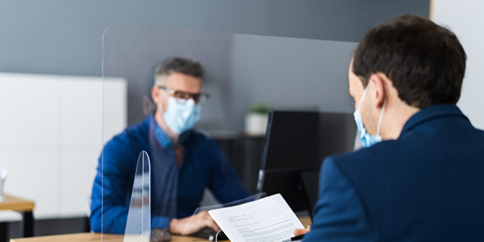 Zwei Männer sitzen mit Maske im Büro mit Plexiglas