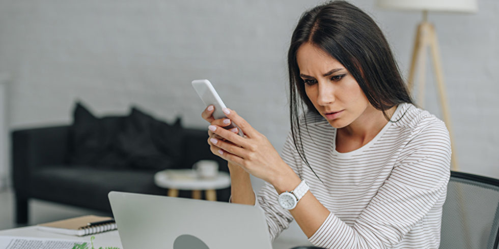 Frau hält Handy in der Hand und schaut auf Laptop