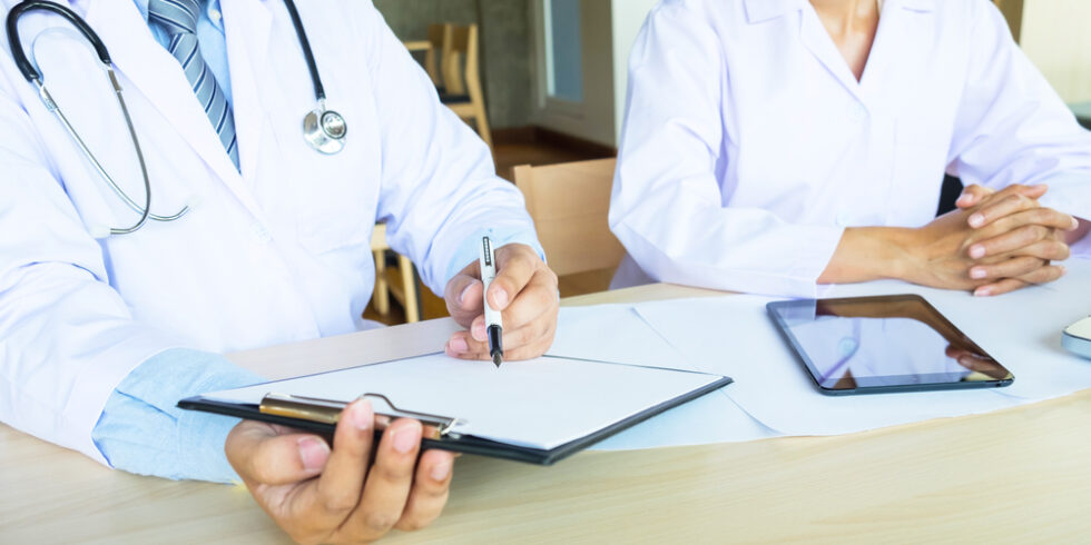 Ärztin und Arzt sitzen mit Schreibutensilien und Tablet an einem Tisch.