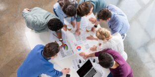 Blick von oben auf eine Gruppe Menschen, die um einen Tisch herumstehen, mit Papieren und Laptop darauf und diskutieren über Aufgaben und Ergebnisse.