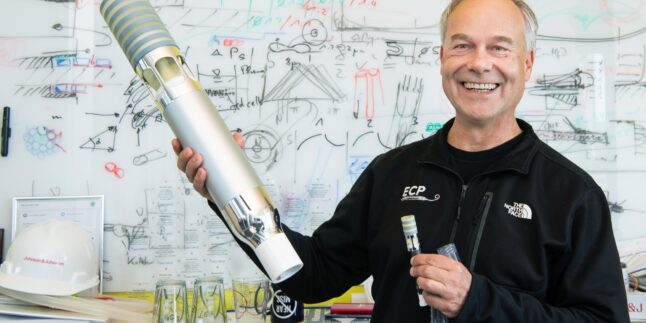 Thorsten Siess steht grinsend vor einem beschriebenen Whiteboard und hält ein technisches Gadget in die Kamera.