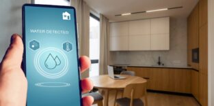 Eine Hand, die ein Smartphone hält, auf der eine App mit Bezug zum Trinkwasser zu sehen ist. Der Blick fällt in eine kleine Küche mit einem Tisch und Sitzgelegenheiten.