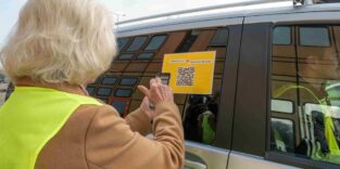 Frau steht vor einem Kleinbus. Auf der Scheibe befindet sich ein Zettel mit einem QR-Code. Die Frau scannt diesen QR-Code mit ihrem Smartphone.