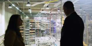 Zwei Personen betrachten durch eine Glasscheibe ein automatisiertes Hochregallager.