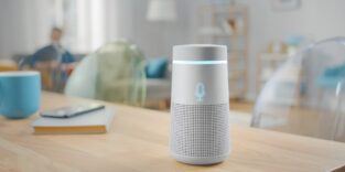 Alexa, Gerät zur Sprachsteuerung, steht auf einem Küchentisch. Daneben liegt ein Smartphone auf einem Buch. Im Hintergrund ist eine Wohnszene zu sehen.