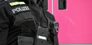 Eine Puppe mit Polizei-Unform steht auf der Digitalmesse Digital X vor einem Containerstand der Deutschen Telekom