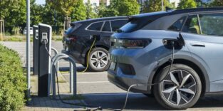 Zwei Elektroautos stehen versetzt hintereinander und sind jeweils an eine Ladesäule angeschlossen. Im Hintergrund ist ein Wohnviertel sichtbar.