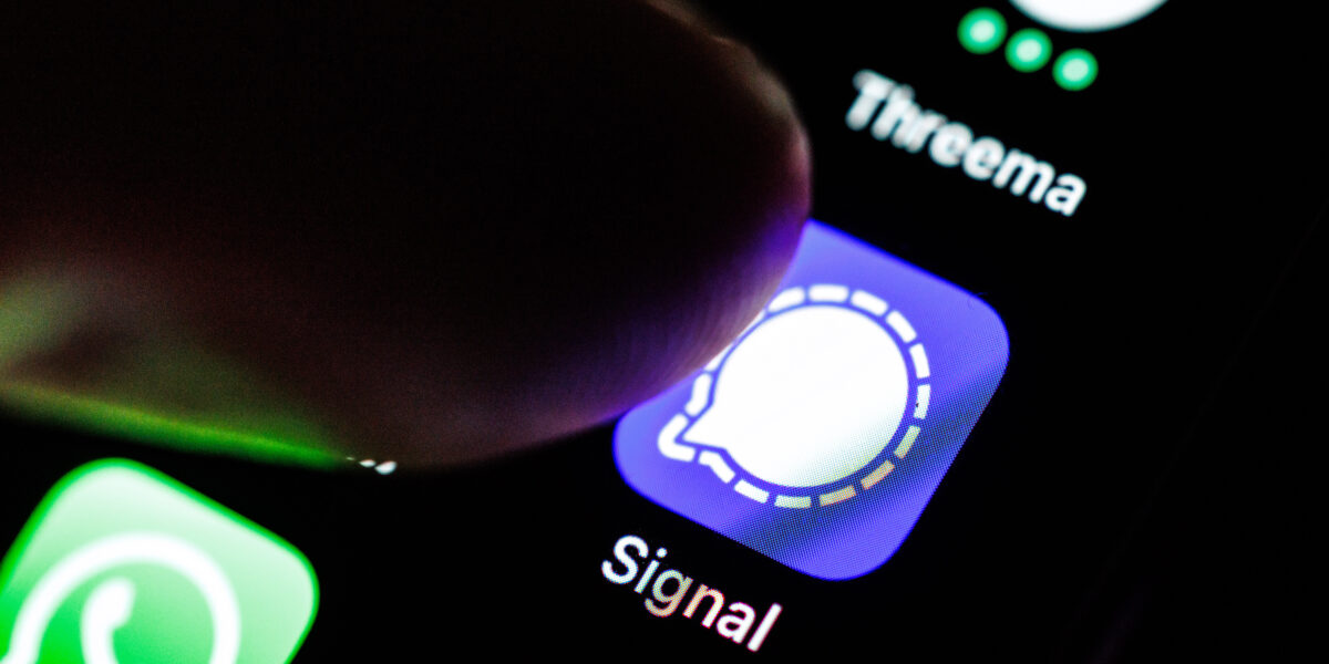 Das Logo der App Signal durch eine Lupe auf dem Display von einem Smartphone zu sehen, während ein Zeigefinger sich dem Symbol der Applikation nähert.