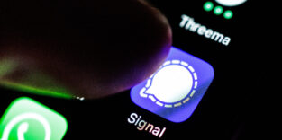 Das Logo der App Signal durch eine Lupe auf dem Display von einem Smartphone zu sehen, während ein Zeigefinger sich dem Symbol der Applikation nähert.