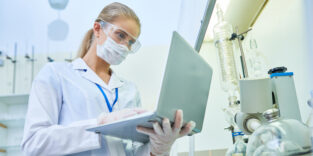 Forscherin im Laborkittel und mit Schutzmaske betrachtet konzentriert einen Laptop in einem Laborumfeld.