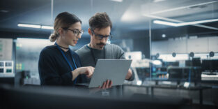 Portrait of Two Creative Young Female and Male Engineers Using Laptop Computer to Analyze and Discuss How to Proceed with the Artificial Intelligence Software. Standing in High Tech Research Office