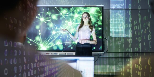 Eine junge Frau steht vor einem großen interaktiven Display und präsentiert ein digitales Netzwerk aus grünen Knoten und Verbindungslinien. Auf ihr und im Raum sind Projektionen von Programmcode und binären Zeichen zu sehen, die Themen wie Informatik, Datenverarbeitung und Künstliche Intelligenz visualisieren.