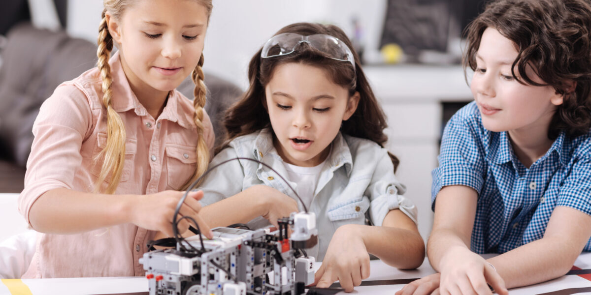 Drei Kinder sitzen an einem Tisch und bauen gemeinsam einen kleinen Roboter aus einem Baukastensystem. Sie konzentrieren sich auf Kabel und Bauteile, während der Roboter auf einer bedruckten Spielfläche steht.