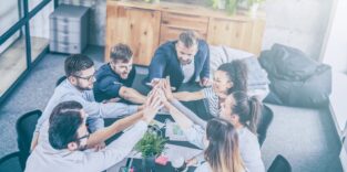 Gruppe sitzt in einem Besprechungsraum um einen Tisch und fasst sich in der Tischmitte an den Händen an.