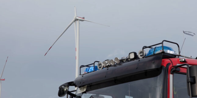 Ein Fahrzeug der Feuerwehr steht vor einem Windrad bei dem ein Flügel beschädigt ist