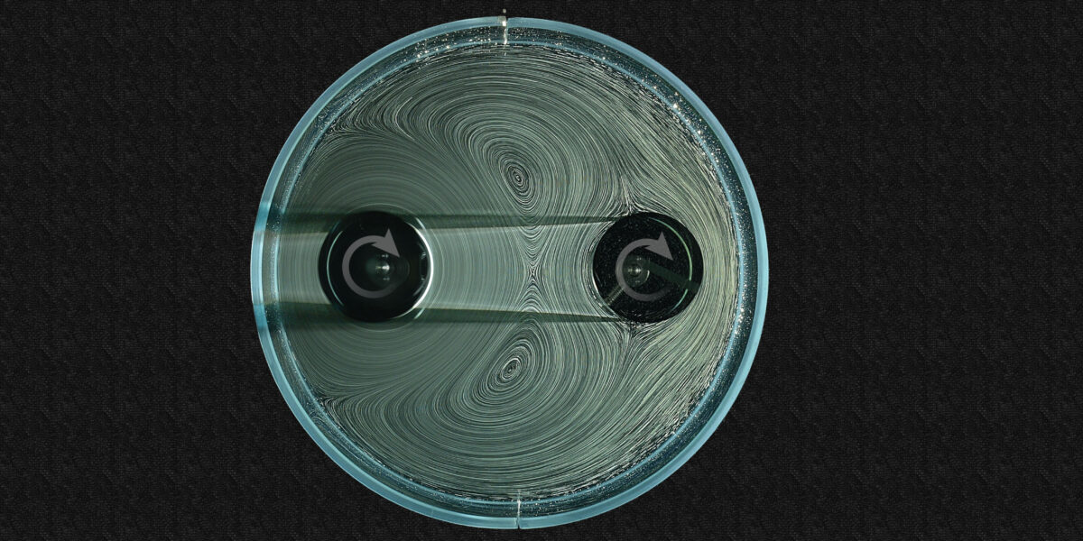 Zwei Spinner in einem kreisförmigen Behälter, umgeben von Flüssigkeit mit Blasen, die helfen, die Strömungen sichtbar zu machen. Der linke Spinner wird aktiv durch einen Motor (nicht abgebildet) angetrieben, während sich der rechte Spinner passiv aufgrund der Strömungen dreht