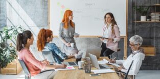 Mehrere Frauen unterschiedlicher Altersgruppen sitzen in einem modernen Besprechungsraum an einem Konferenztisch und diskutieren. Zwei Frauen stehen vor einem Whiteboard mit einer skizzierten Organisationsstruktur und präsentieren Inhalte, während andere Notizen machen oder an Laptops arbeiten. Die Szene vermittelt Teamarbeit, Austausch und Weiterbildung im beruflichen Kontext.