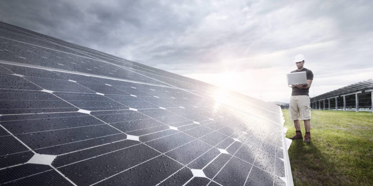 Nahaufnahme eines Solarmoduls in einer Freiflächenanlage, auf dessen dunkler Oberfläche zahlreiche Wassertropfen zu sehen sind. Im Hintergrund steht ein Mann mit Schutzhelm und Laptop zwischen den Modulreihen, während die Sonne durch eine bewölkte Wolkendecke bricht.