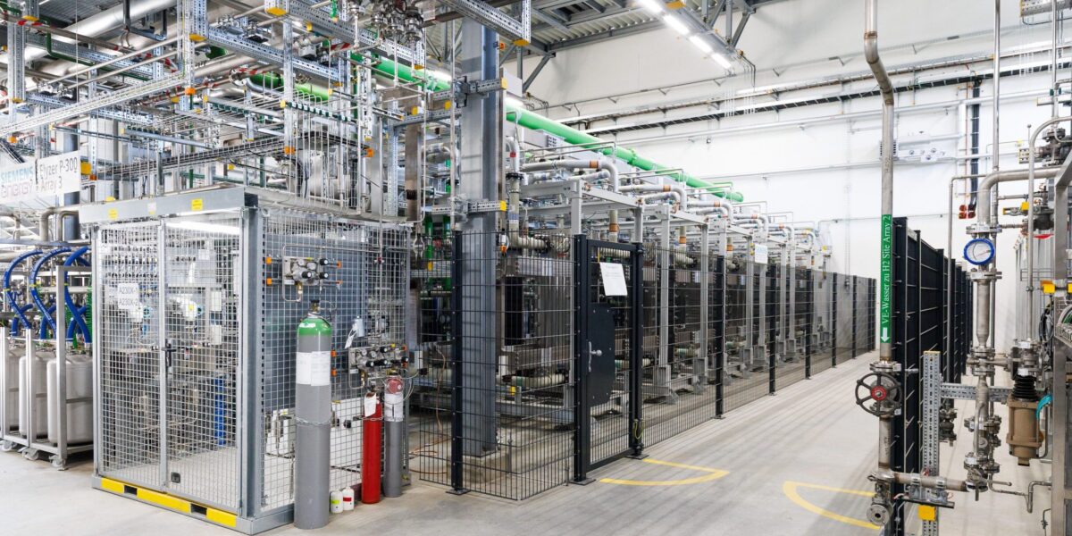 Blick in die Elektrolyseurhalle von BASF in Ludwigshafen: Hinter Schutzgittern stehen mehrere PEM-Elektrolyseur-Module, verbunden durch ein Netz aus Rohrleitungen und Versorgungsleitungen. Mit 54 MW Leistung ist es derzeit die größte PEM-Elektrolyseanlage in Deutschland.