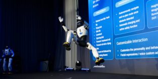 Ein humanoider Roboter springt bei einer Präsentation auf einer Bühne. Im Hintergrund steht ein weiterer humanoider Roboter .