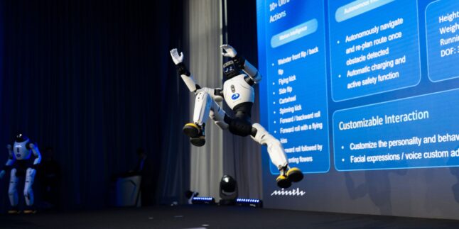 Ein humanoider Roboter springt bei einer Präsentation auf einer Bühne. Im Hintergrund steht ein weiterer humanoider Roboter .