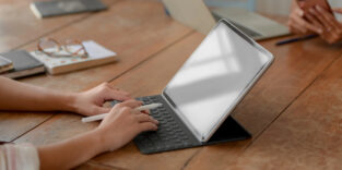 Person arbeitet an einem Tablet mit Tastatur und Stift an einem Holztisch, offenbar bei einer handschriftlichen Aufgabe oder digitalen Notizen.