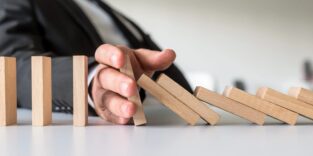 Weitwinkelaufnahme eines Unternehmensmediators, der in einem Konzeptbild einer Unternehmenskrise einen fallenden Dominostein aus Holz mit der Hand stoppt.