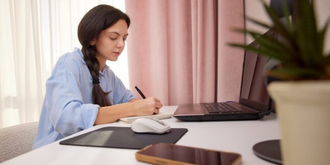 Frau, die in ein Notizbuch schreibt, während sie am Schreibtisch mit einem Laptop arbeitet oder lern.