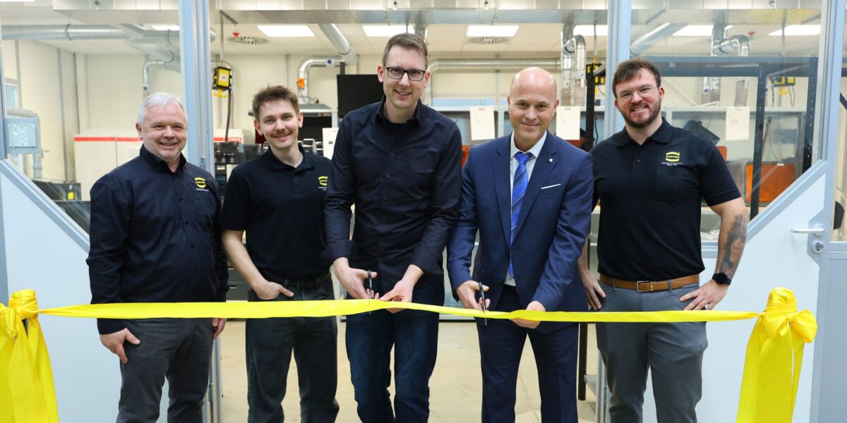 Fünf Männer stehen nebeneinander vor dem Eingang eines Fertigungsraums und durchschneiden ein gelbes Band zur Eröffnung des Kompetenzzentrums. Drei tragen schwarze Hemden mit Harting-Logo, einer ein dunkelblaues Hemd, einer einen blauen Anzug mit Krawatte. Im Hintergrund sind 3D-Drucker und technische Anlagen zu erkennen.