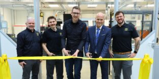 Fünf Männer stehen nebeneinander vor dem Eingang eines Fertigungsraums und durchschneiden ein gelbes Band zur Eröffnung des Kompetenzzentrums. Drei tragen schwarze Hemden mit Harting-Logo, einer ein dunkelblaues Hemd, einer einen blauen Anzug mit Krawatte. Im Hintergrund sind 3D-Drucker und technische Anlagen zu erkennen.