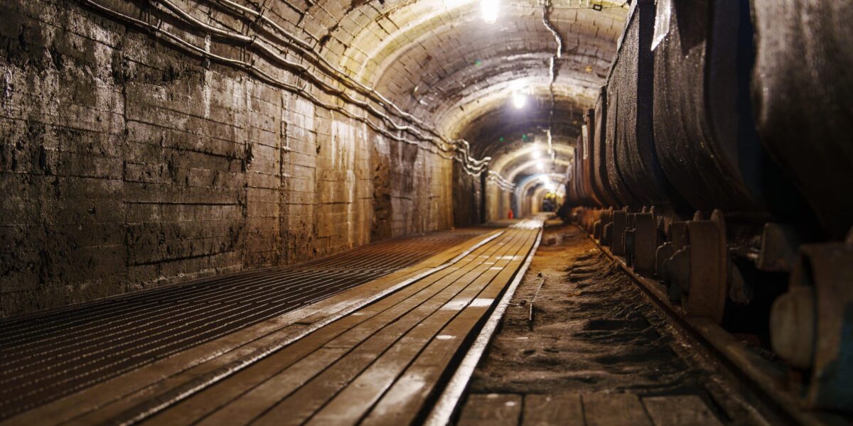 Blick in einen beleuchteten Bergwerksstollen mit Gleisen und Holzbohlen, der in die Tiefe führt.