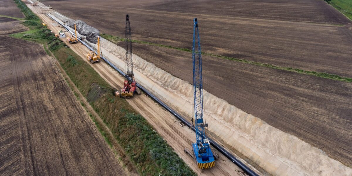 au einer Gasleitung: Ein Seitenbaumleger hebt ein großes Stahlrohr in einen offenen Leitungsgraben, im Hintergrund weitere Baumaschinen und Erdaushub.