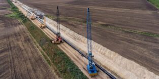 au einer Gasleitung: Ein Seitenbaumleger hebt ein großes Stahlrohr in einen offenen Leitungsgraben, im Hintergrund weitere Baumaschinen und Erdaushub.