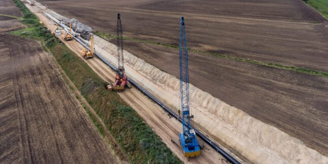 au einer Gasleitung: Ein Seitenbaumleger hebt ein großes Stahlrohr in einen offenen Leitungsgraben, im Hintergrund weitere Baumaschinen und Erdaushub.