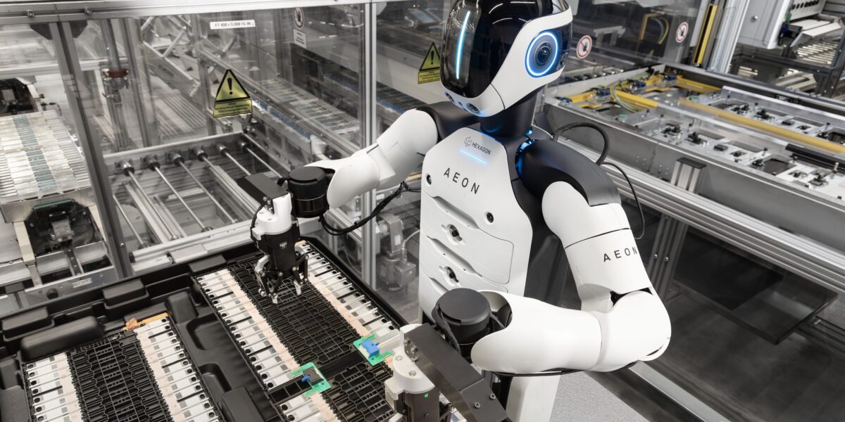 Ein humanoider Roboter mit weißer Brust und weißen Armen bei der Handhabung kleinerer Automobilkomponenten in einer automatisierten Anlage.