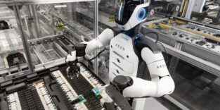 Ein humanoider Roboter mit weißer Brust und weißen Armen bei der Handhabung kleinerer Automobilkomponenten in einer automatisierten Anlage.