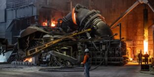 Arbeiter vor einem gekippten Elektrolichtbogenofen mit glühender Schlacke und orangefarbenem Feuerschein in einer Stahlwerkshalle