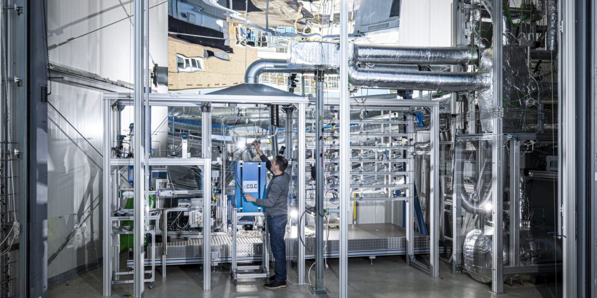 Ein Forscher steht vor der mehrstöckigen NECOC-Versuchsanlage am KIT. Zu sehen sind Rohrleitungen, ein zentral angeordneter Ofen mit Blasensäulenreaktor sowie Gasaufbereitungs- und Filtersysteme in einer Laborhalle.